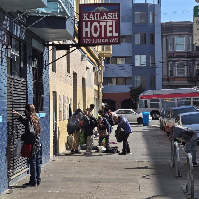 A group of people gather on the sidewalk near Kailash Hotel, 179 Julian Ave. Some are interacting and others are leaning against the wall. A bus is visible on the street.
