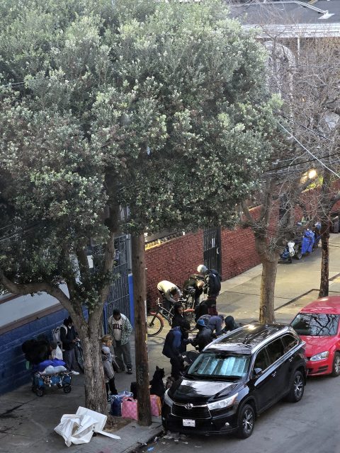 A group of people gathered on a sidewalk near a parked black SUV, with bicycles and trees in the background.