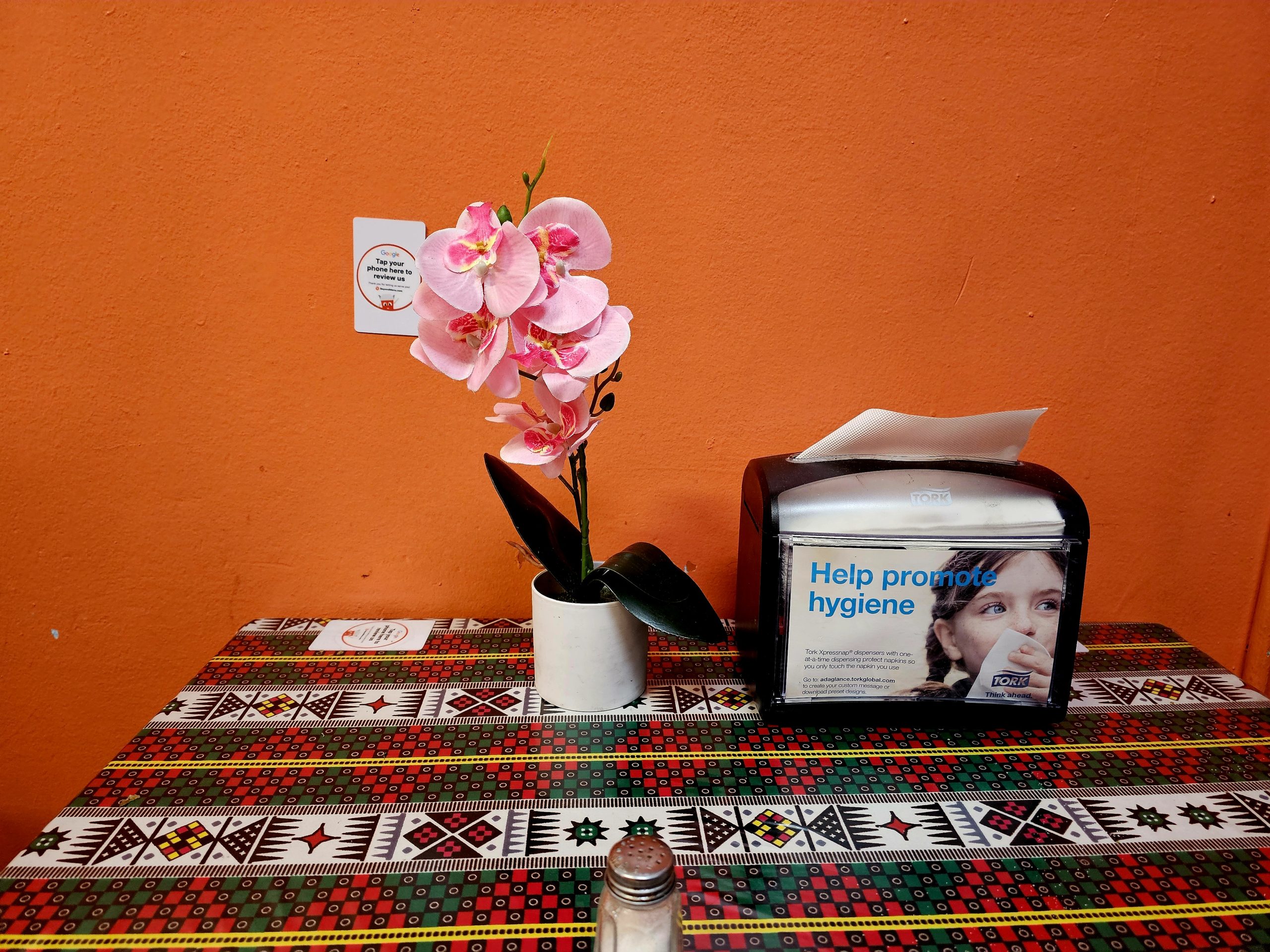 Orchid in a white pot next to a napkin holder on a patterned table with an orange wall backdrop.