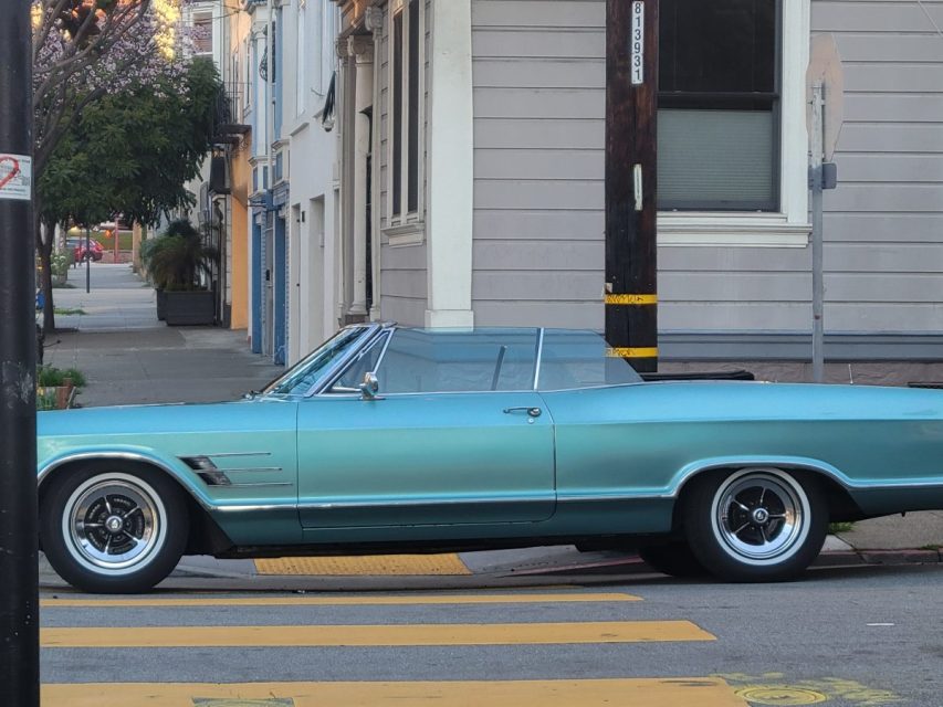 Teal vintage convertible car parked on a city street beside a crosswalk. Buildings and a tree are in the background.