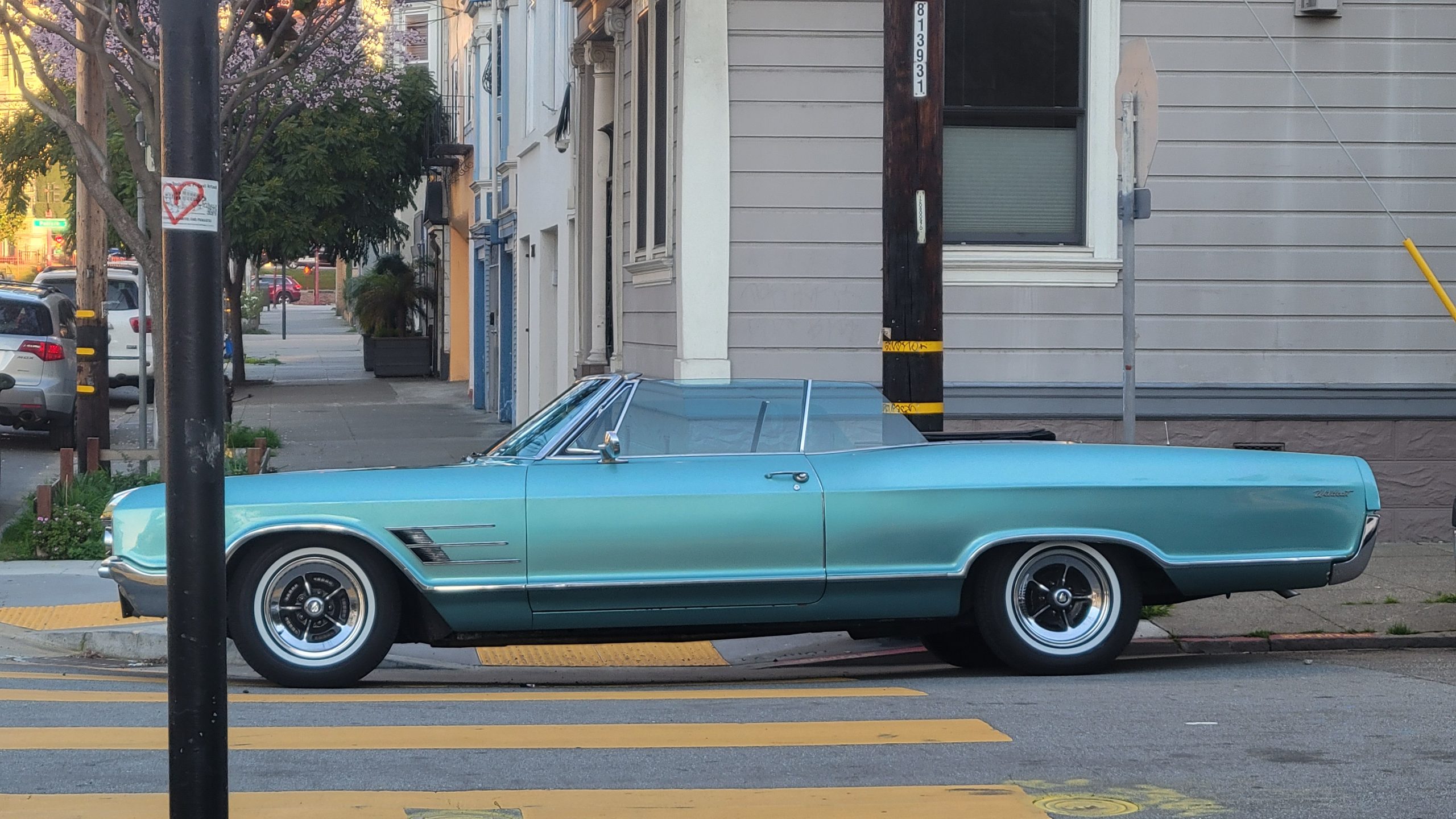A vintage turquoise convertible car is parked on a street corner beside a gray building. Trees and other vehicles are visible in the background.
