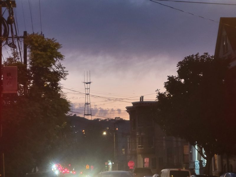 City street at dusk with cars and traffic lights; distant hills and a tower are silhouetted against a blue and purple sky.