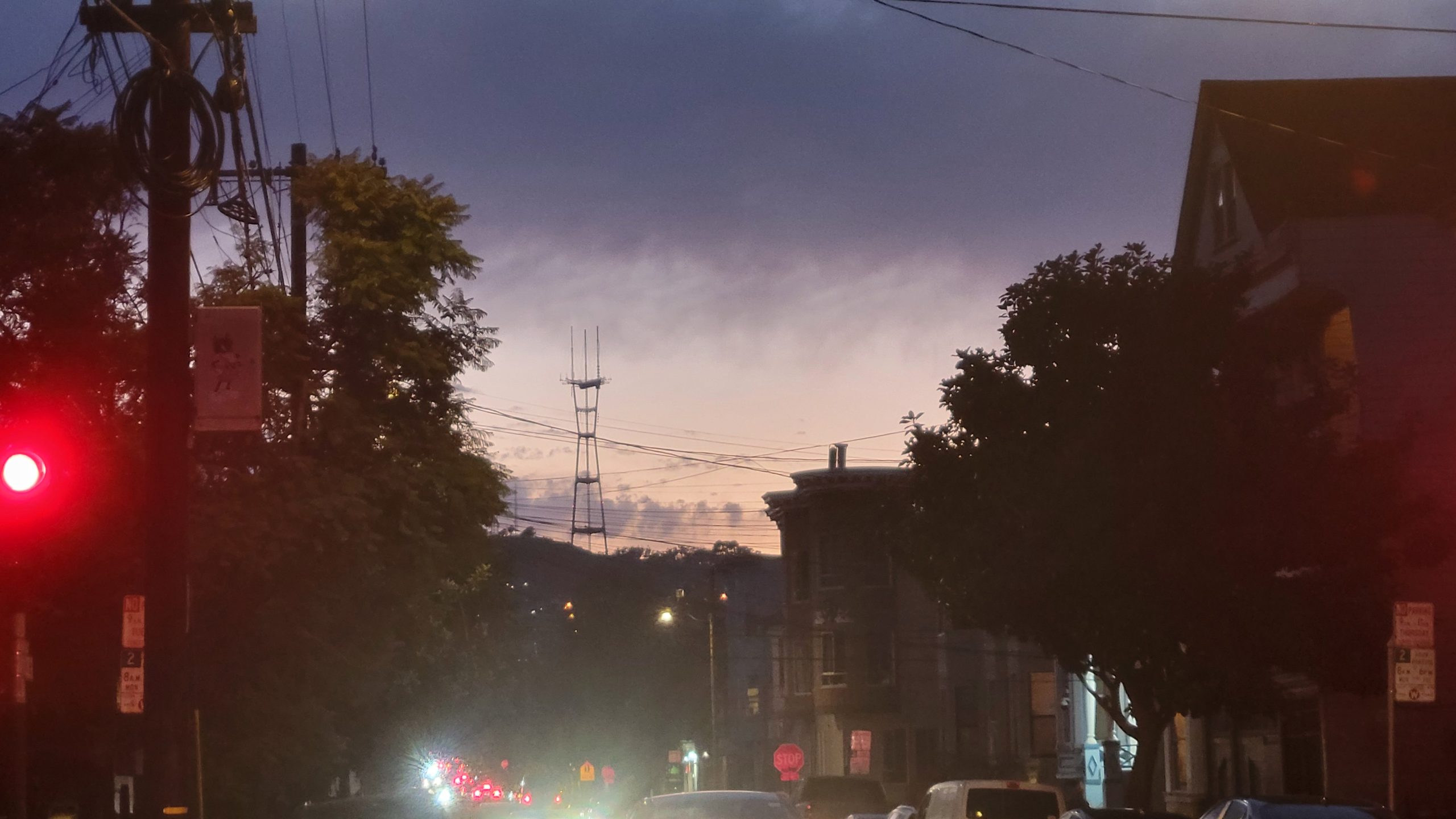 City street at dusk with cars and traffic lights; distant hills and a tower are silhouetted against a blue and purple sky.