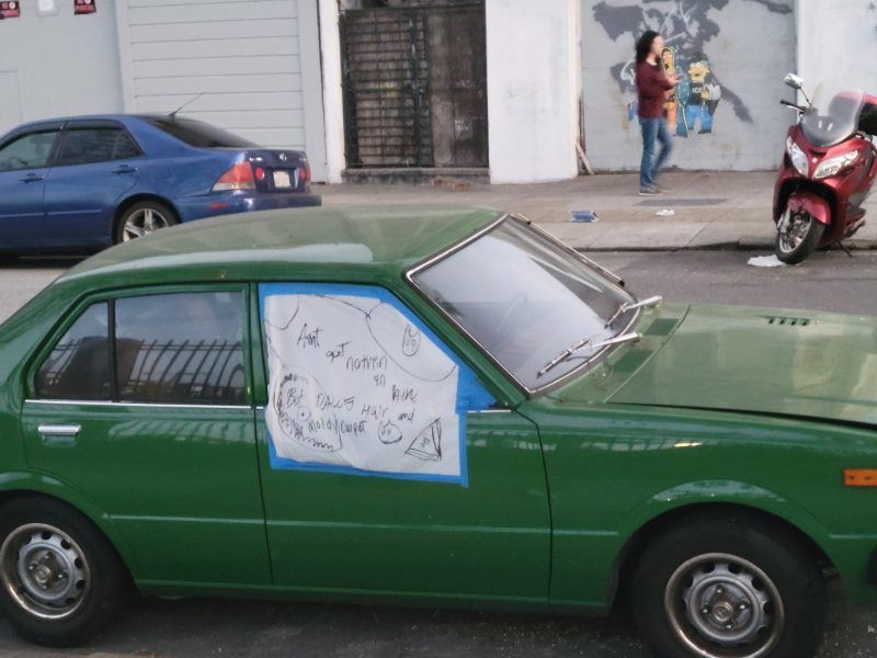 Green vintage car parked on the street with handwritten paper notes taped to windows. A person is in the background near a wall with a mural. A blue car and red scooter are also visible.