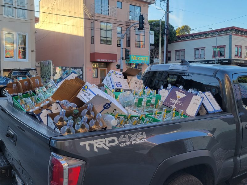 Pickup truck bed filled with recyclable cardboard boxes and plastic bottles parked on a city street.