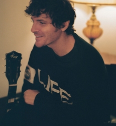 Person sitting in a dimly lit room, smiling and looking to the side, with a guitar headstock visible in the foreground and a lamp in the background.
