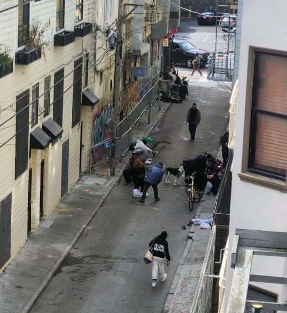 A narrow urban alleyway with people walking, a few sitting on the ground with belongings, a parked bicycle, and buildings with graffiti and utility wires.