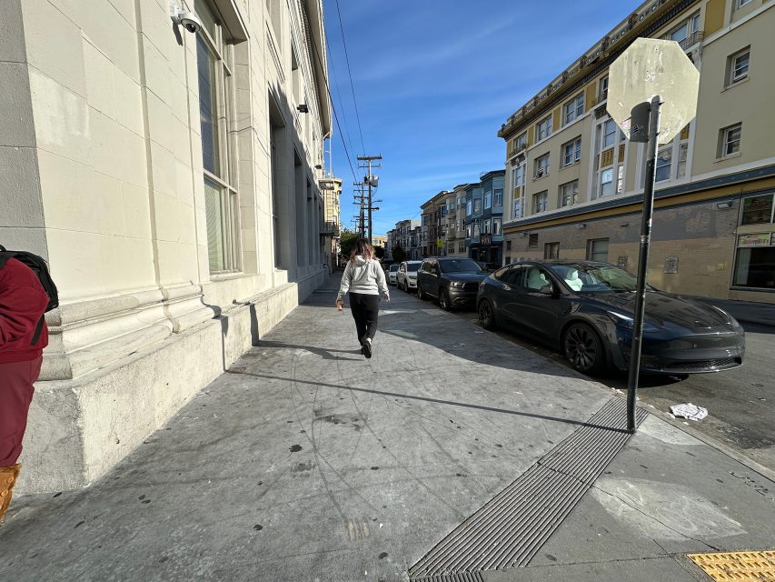 Person walking on a city sidewalk lined with parked cars and buildings under a clear blue sky.