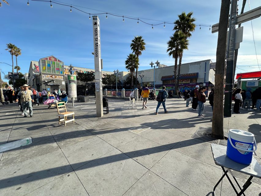 Urban street scene with people walking and gathering near colorful buildings and palm trees. A blue cooler and chair are in the foreground, with string lights overhead.