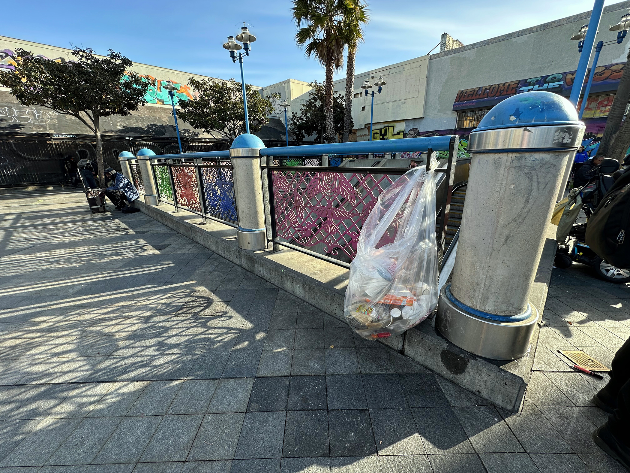 A public area with a metal fence, a trash bag hanging on it, palm trees, and buildings with murals in the background.