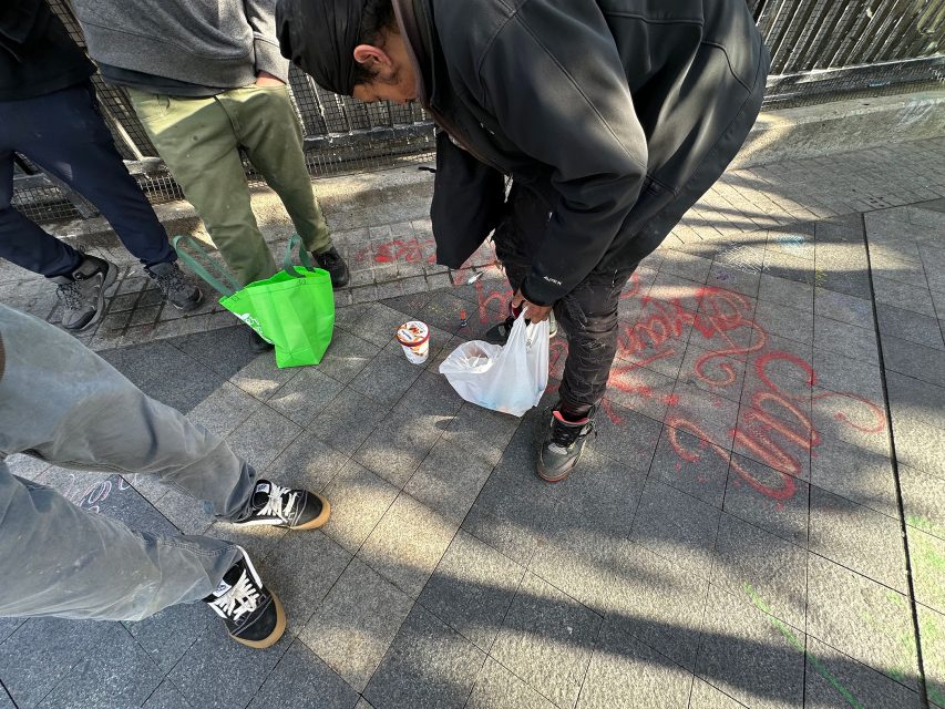 People standing on a paved area. A person is bending over, holding a plastic bag, with a cup on the ground. Green bag nearby. Sidewalk has red graffiti and shadow patterns.
