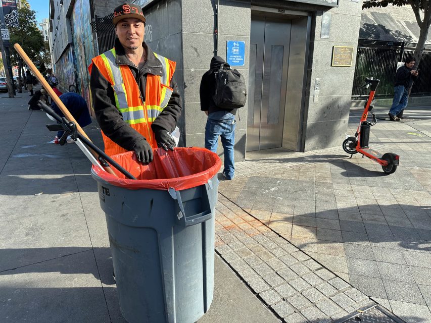 A person in a high-visibility vest stands beside a trash can holding a broom and dustpan. An electric scooter is on the sidewalk, and an elevator is in the background.