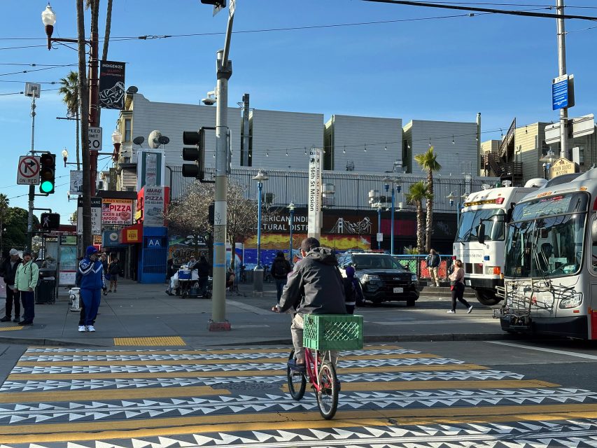 A cyclist on a patterned crosswalk at an urban intersection, with buses, pedestrians, and various storefronts in the background on a clear day.
