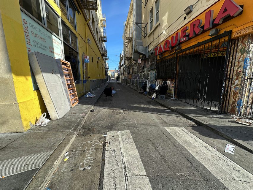 Narrow urban alley with graffiti on walls, a "Taqueria" sign, two people sitting against a wall, and scattered debris.