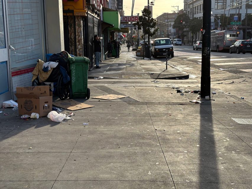 Urban street scene with litter and cardboard on the sidewalk, next to a green trash bin. People stand near storefronts and a liquor store sign. Vehicles and a bus are visible on the road.