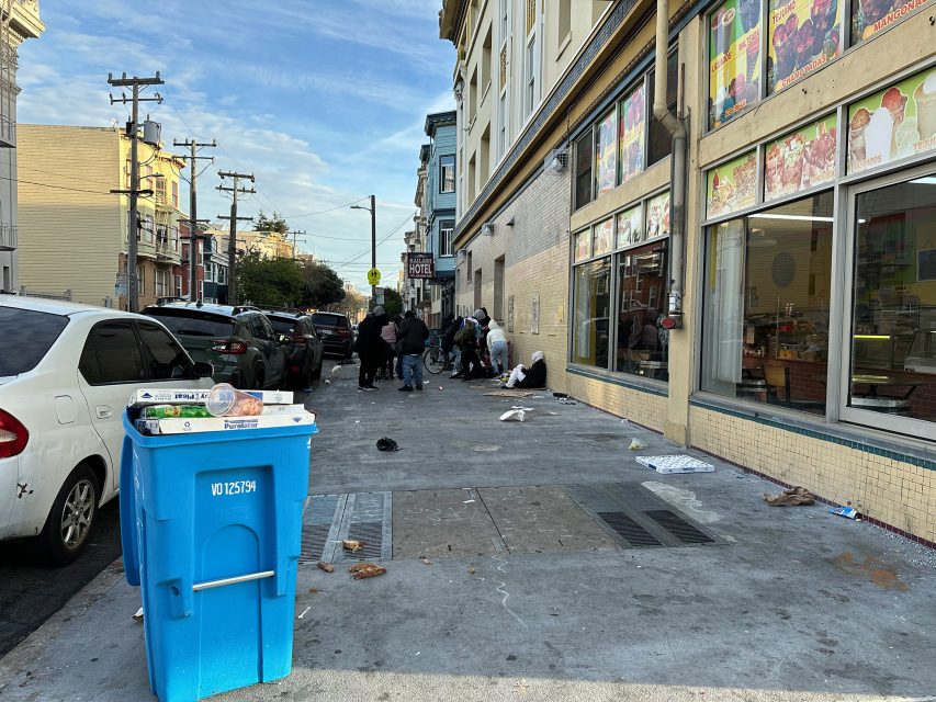 Urban street scene with people gathered near a storefront, surrounded by litter. A blue recycling bin and parked cars are visible. Buildings and power lines line the street.