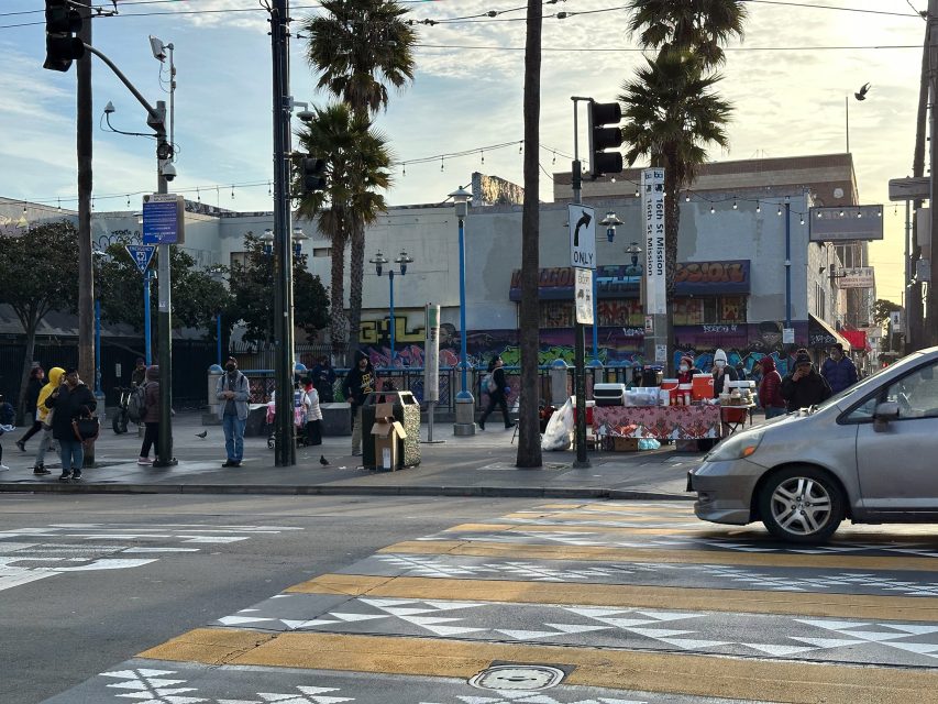 Street scene with pedestrians crossing and browsing market stalls. Palm trees, graffiti, and buildings are in the background. A car is driving in the foreground.