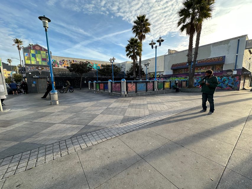A person stands in a plaza with colorful murals, palm trees, and nearby people. The sky is partly cloudy, and the area is paved with concrete tiles.