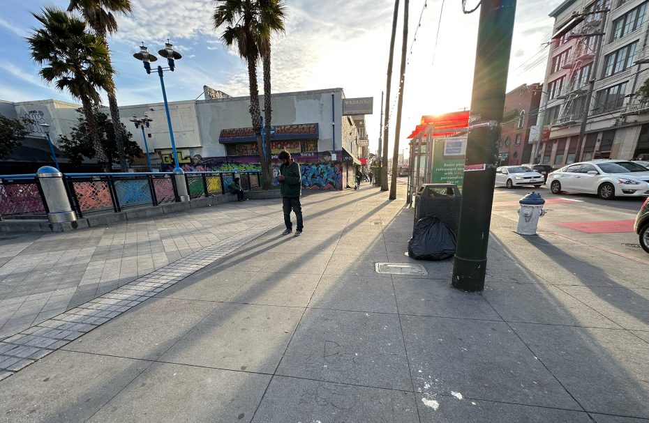 A person walks along a sunny urban street lined with palm trees. There are graffiti-covered buildings and parked cars.