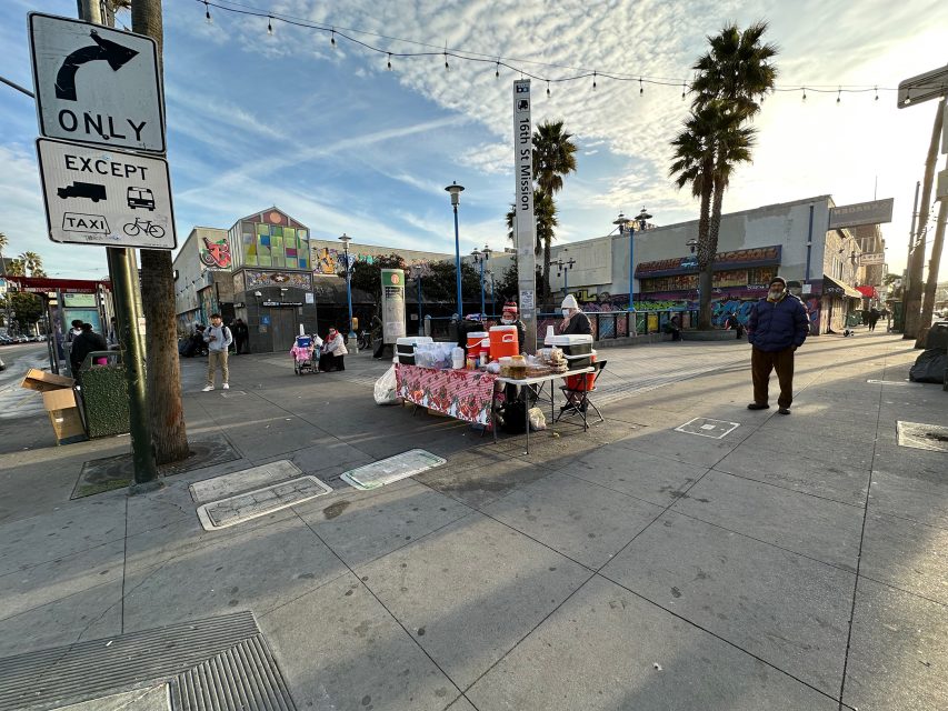Street scene with food stall under a tent, colorful mural wall in background, pedestrian crossing signs, and palm trees. Cloudy sky with string lights above. People walking and standing nearby.