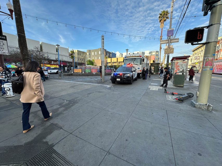 A police car with flashing lights is parked on a city street near a bus stop. Two officers are interacting with a person near the vehicle. Pedestrians and an electric scooter are nearby.