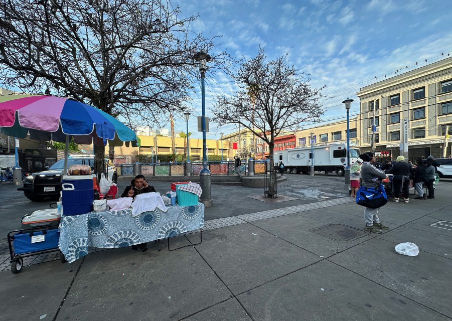 Street scene with a vendor under a colorful umbrella, selling items on a table. Trees without leaves and surrounding buildings are visible under a partly cloudy sky. People walk nearby.