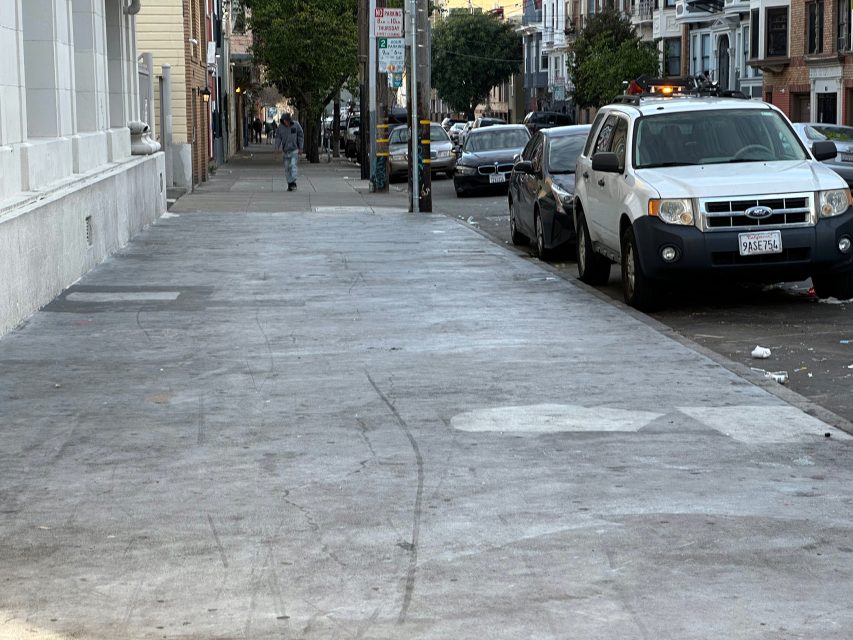 A quiet urban street with parked cars along the side. A lone pedestrian walks away in the distance next to a row of buildings.
