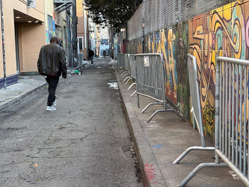 An alley with graffiti on the wall, metal barricades, scattered trash, and two people walking away.
