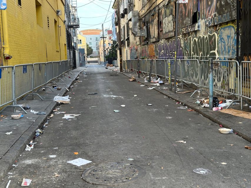 An urban alley with graffiti-covered walls and metal barriers. Litter, including papers and plastic items, is scattered on the ground. A fire escape and utility wires are visible above.