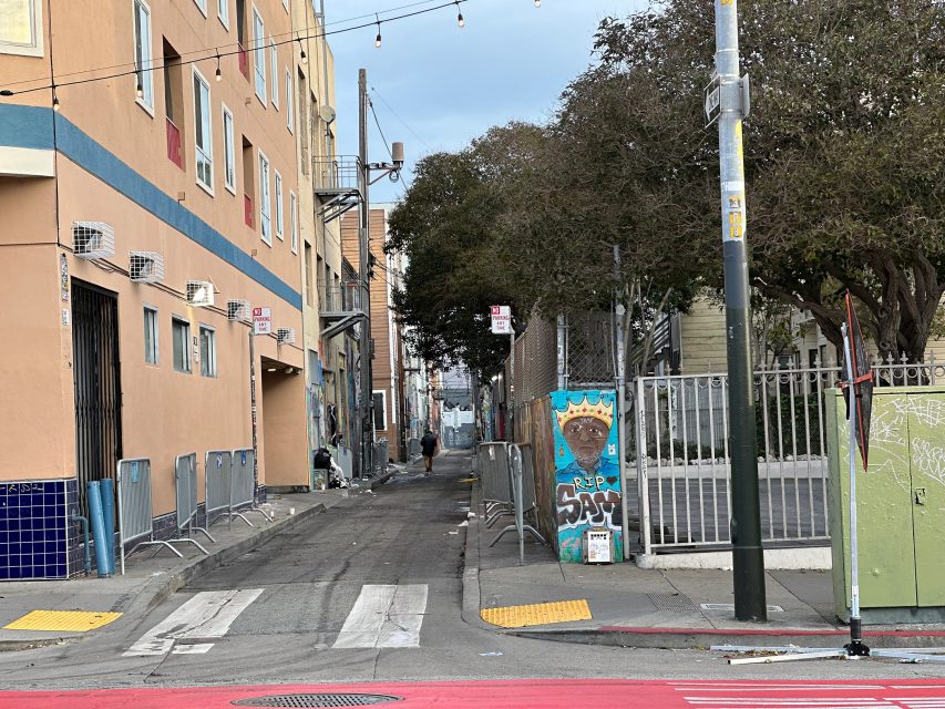 Narrow urban alley with a colorful mural and graffiti on walls, lined with row of buildings and trees, streetlights, and overhead string lights, leading to a distant figure.