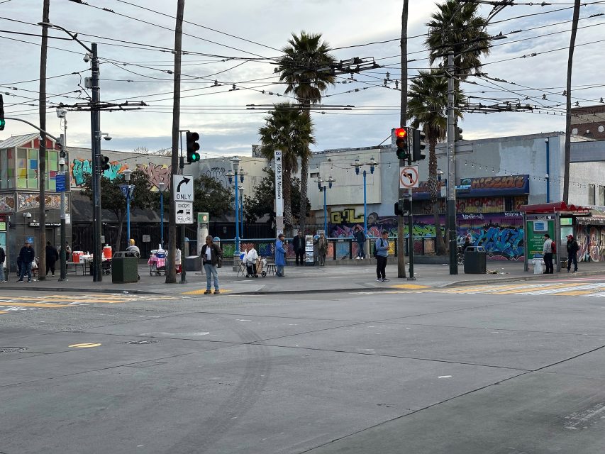 Street scene with people at an intersection, surrounded by palm trees and buildings with graffiti. Traffic signals are visible, and the sky is overcast.