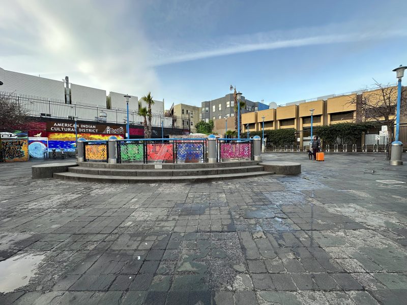 Public square with colorful murals on a circular platform in front of a building labeled "American Indian Cultural District." Overcast sky and wet ground.