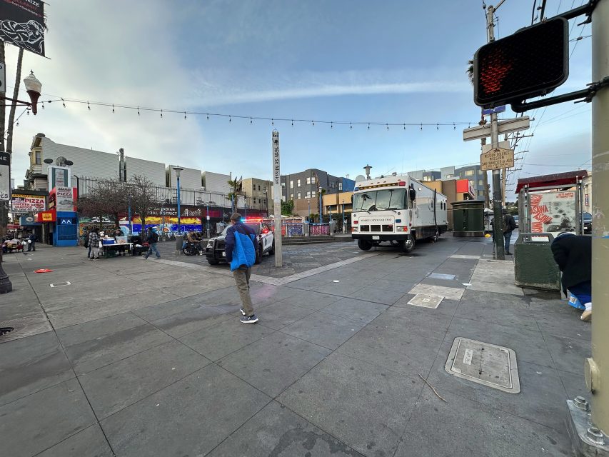 Street scene with a person crossing a crosswalk; police car and truck nearby. Buildings, store signs, and a traffic light are visible in the background.