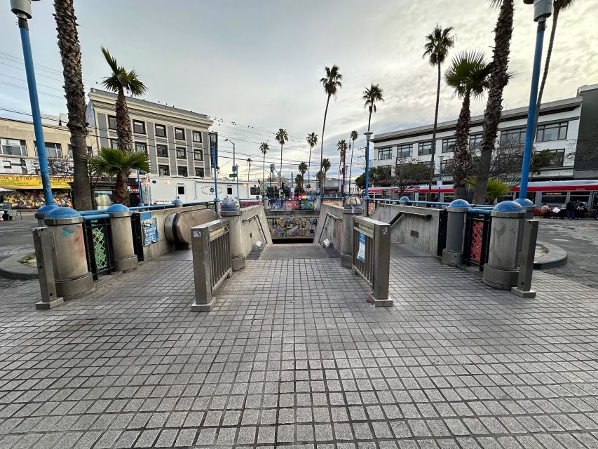 Entrance to an underground metro station in a plaza with palm trees, surrounded by buildings and people in the background.
