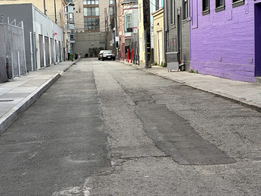 A narrow urban alley with cracked pavement, flanked by buildings on both sides. A car is parked at the far end. One building on the right is painted purple.