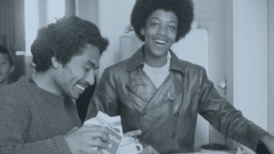 Two people smiling in a kitchen, one holding a cereal box and pouring, the other wearing a leather jacket with an afro hairstyle. A third person is partially visible in the background.