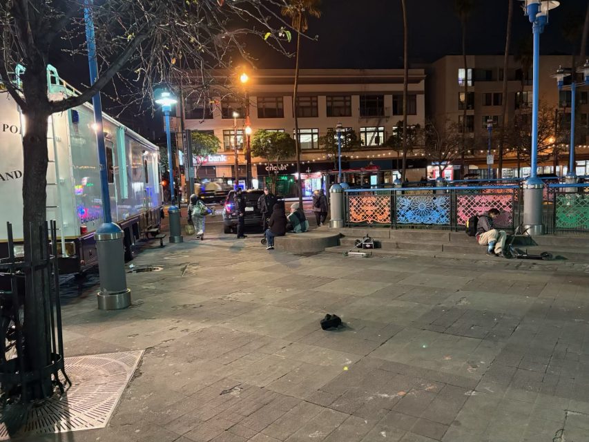 Urban square at night with a few people sitting and lying on benches and pavement. A police vehicle and a lit-up building are in the background. Scattered items are on the ground.