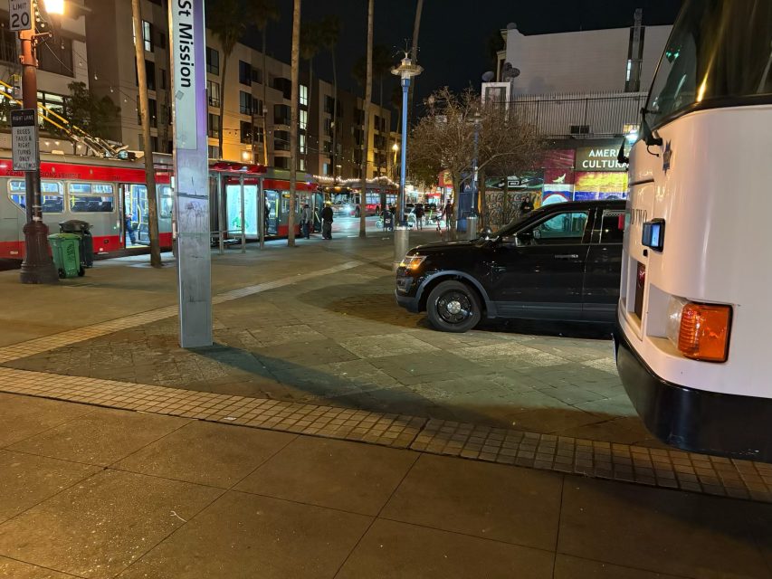Street view at night with buses and an SUV parked near a sidewalk. A row of buildings is illuminated in the background.