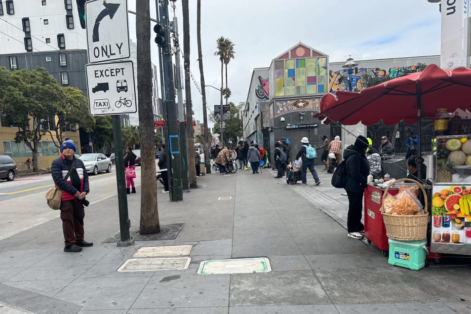 Street scene with people walking and a fruit stand. A person stands by a street pole, wearing a blue beanie. Buildings with murals are in the background.