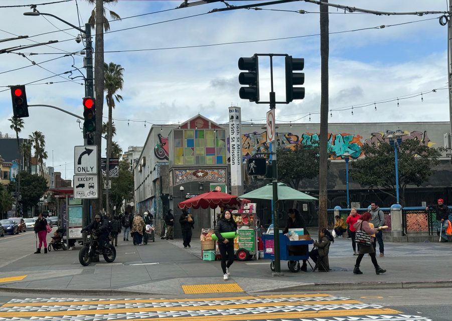 Street scene with people walking, market stalls, traffic lights, palm trees, and a building with colorful mural.