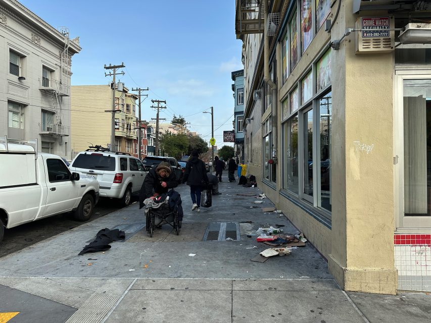 Street scene with people on a sidewalk, some seated and others walking. Vehicles are parked on the left, and the sidewalk has scattered trash. Buildings line the street.