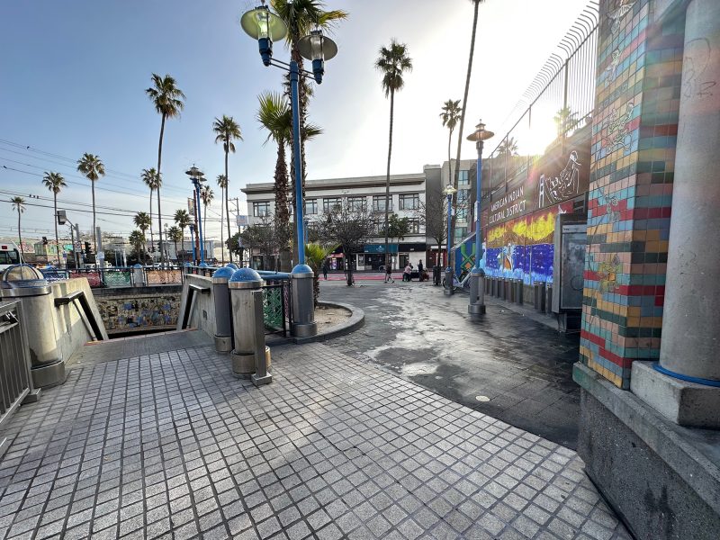 Urban scene with palm trees, a tiled walkway, and a colorful mural on the right. People and a train are visible in the background under a clear sky.
