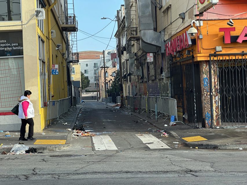 A person with a pink bag stands at a crosswalk next to a trash-strewn alley bordered by a yellow and an orange building.