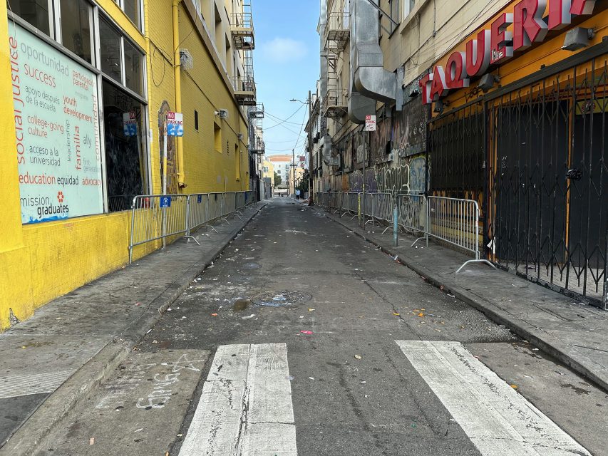 A narrow urban alley with graffiti-covered walls, metal barricades along the sides, and a faded crosswalk in the foreground. Trash is scattered on the asphalt.