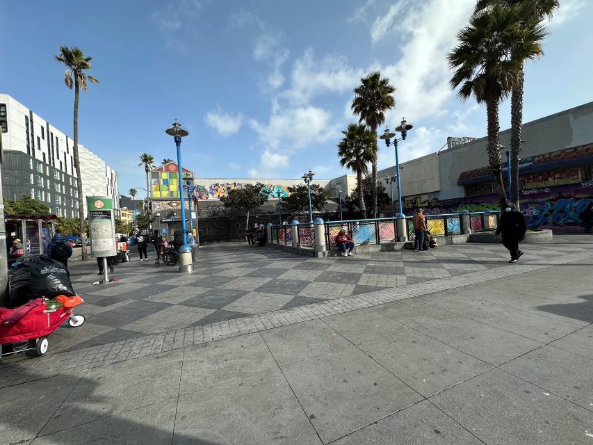 A public square with checkered pavement, palm trees, and graffiti-covered walls. People are walking and sitting. A cart with bags is in the foreground.