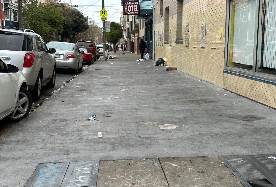 A street with scattered litter, parked cars on the left, and a beige brick building on the right. A hotel sign is visible in the background.