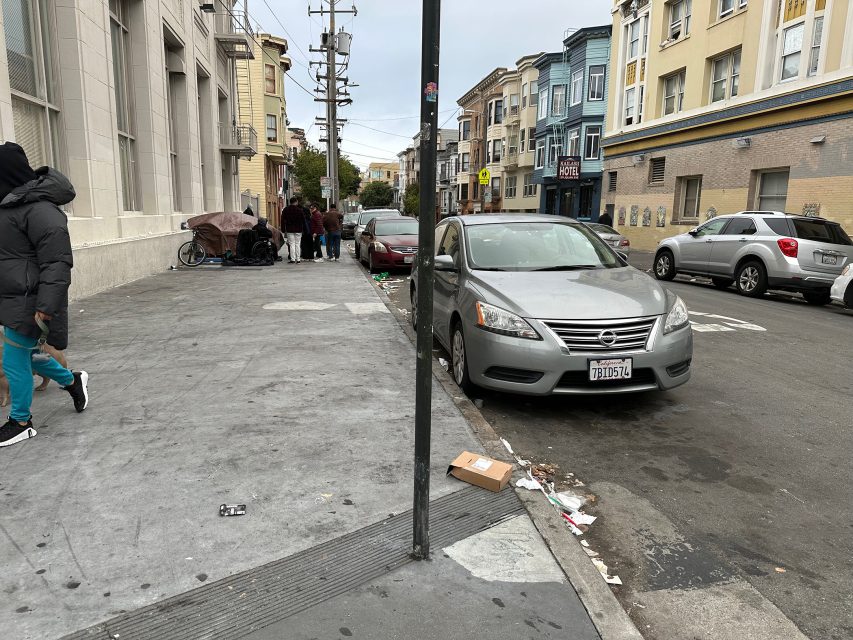 Urban street with parked cars, people walking, and a group gathered near a building. Trash is scattered on the sidewalk, and colorful buildings line the street.