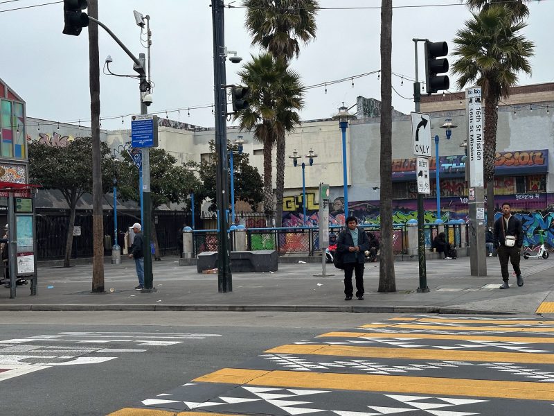 Urban street scene with crosswalk, graffiti-covered walls, palm trees, and a few people walking. Overcast sky and various street signs present.