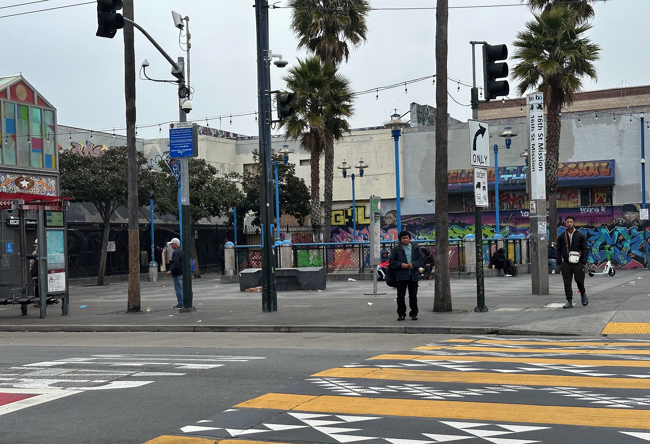 Urban street scene with crosswalk, graffiti-covered walls, palm trees, and a few people walking. Overcast sky and various street signs present.