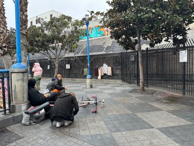 People sitting near a partially dismantled bicycle in a paved area with trees. Clothing hangs on a fence nearby, and a mural is visible in the background.